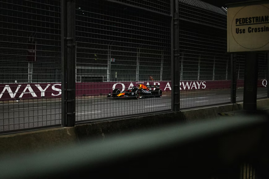 Oracle Red Bull Racing Formula 1 car photographed through a security fence at a night street circuit, with Qatar Airways barrier signage visible
